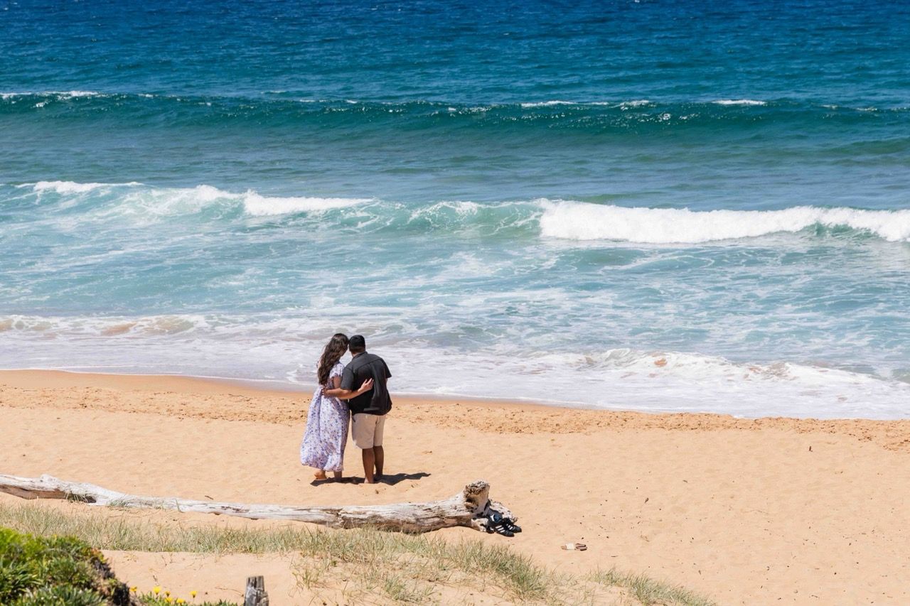 The couple hold each other on the sand with waves behind them