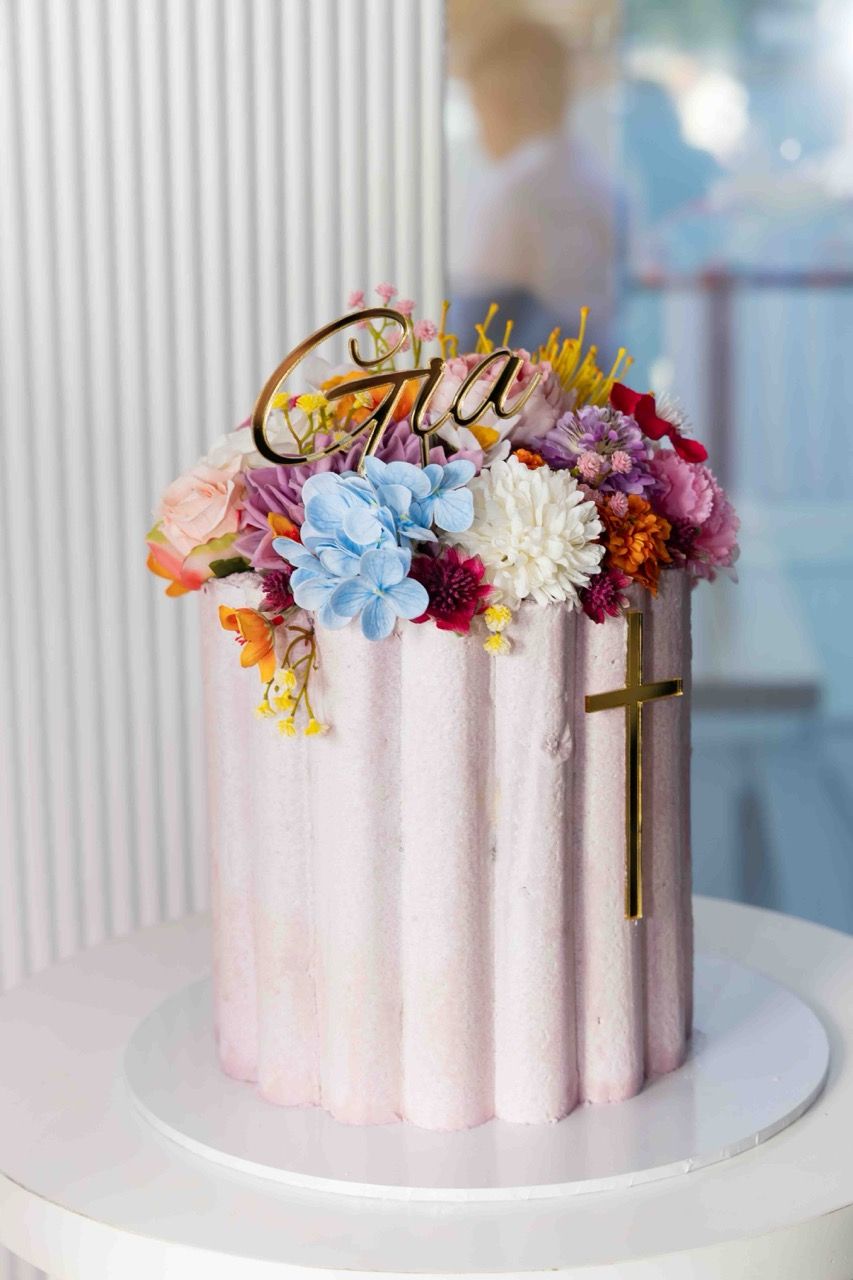 A pink christening cake with flowers and a gold name topper