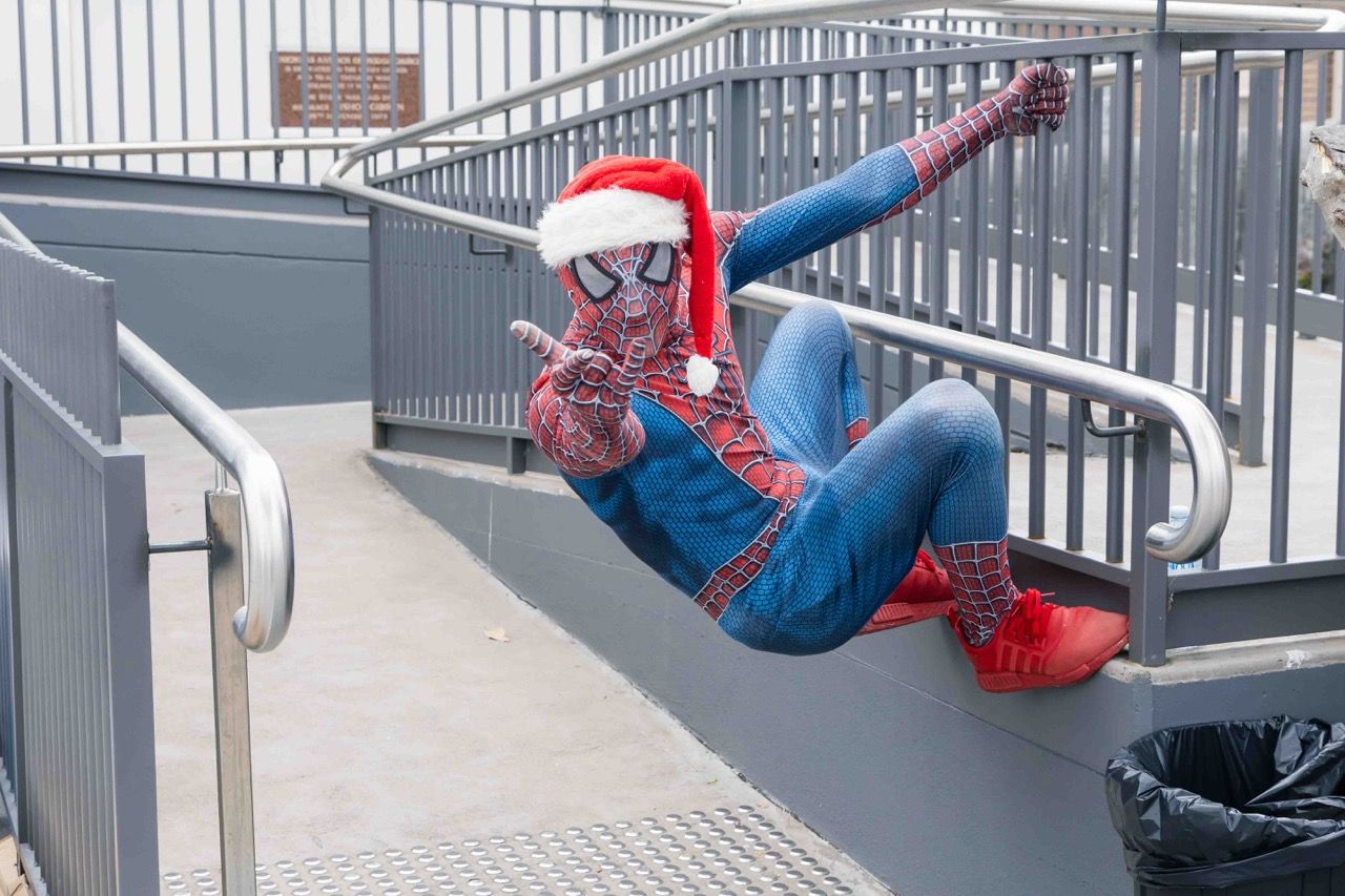 Spider-Man in a Santa hat clings to a stair railing