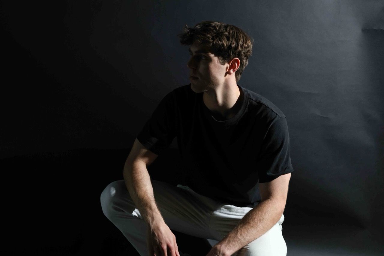 A seated studio portrait in a black t-shirt and white trousers